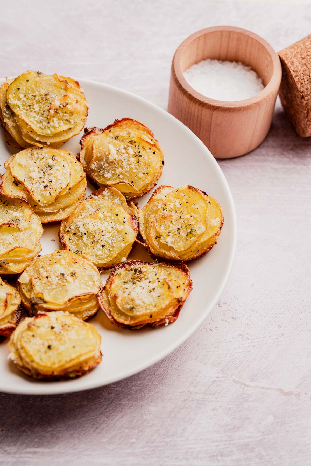 A plate of stacked, baked muffin tin potatoes seasoned with herbs and salt, with a small wooden container of coarse salt in the background.