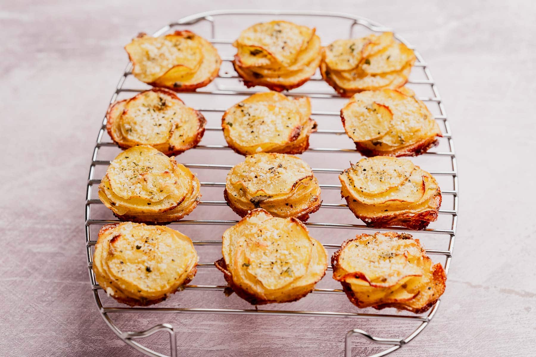 Twelve stacks of thinly sliced, baked muffin tin potatoes with herbs and cheese are arranged on a round cooling rack.