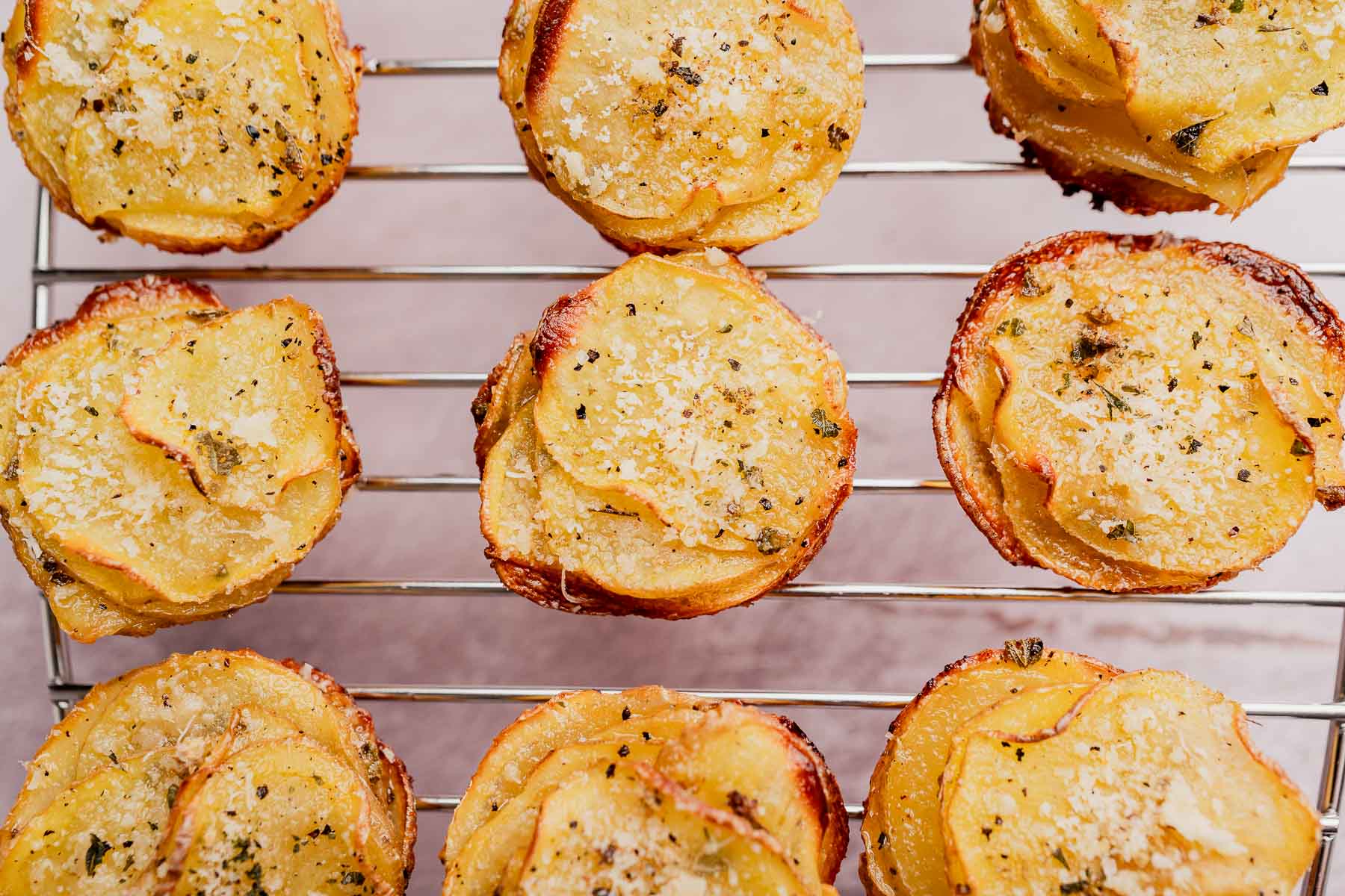 Slices of roasted potatoes topped with herbs and grated cheese are arranged in clusters, just like muffin tin potatoes, on a metal cooling rack.