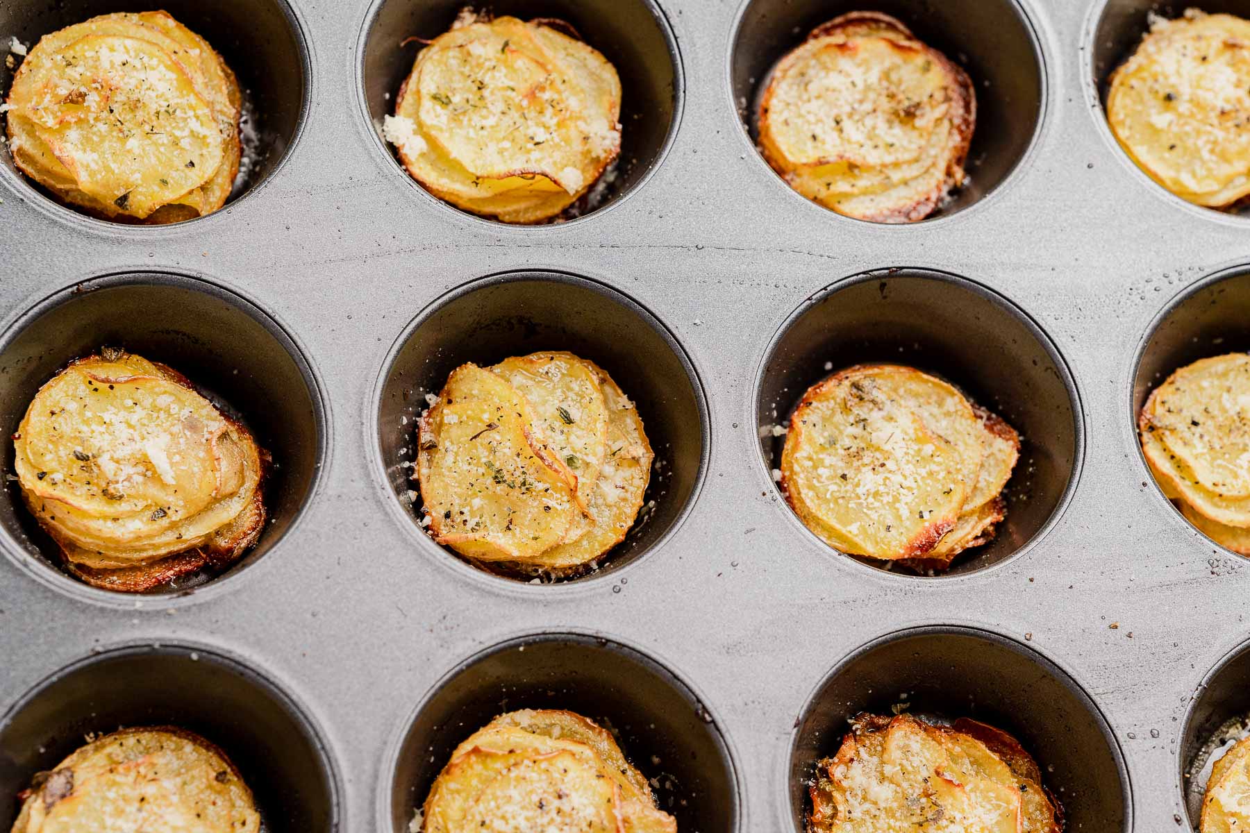 A muffin tin displays several cups of muffin tin potatoes—thinly sliced, baked stacks seasoned generously with herbs for a crispy, flavorful treat.