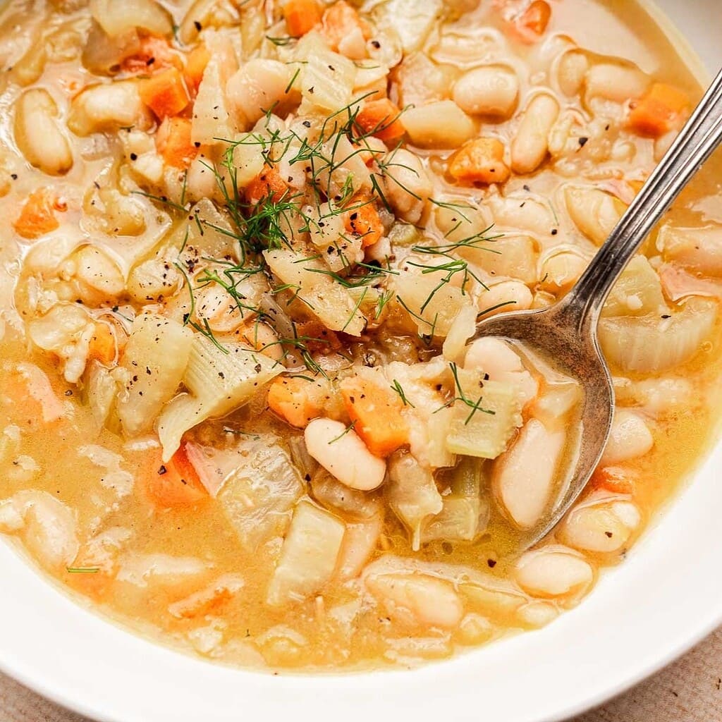A bowl of Tuscan white bean soup with carrots, onions, and fresh herbs is served with a spoon.