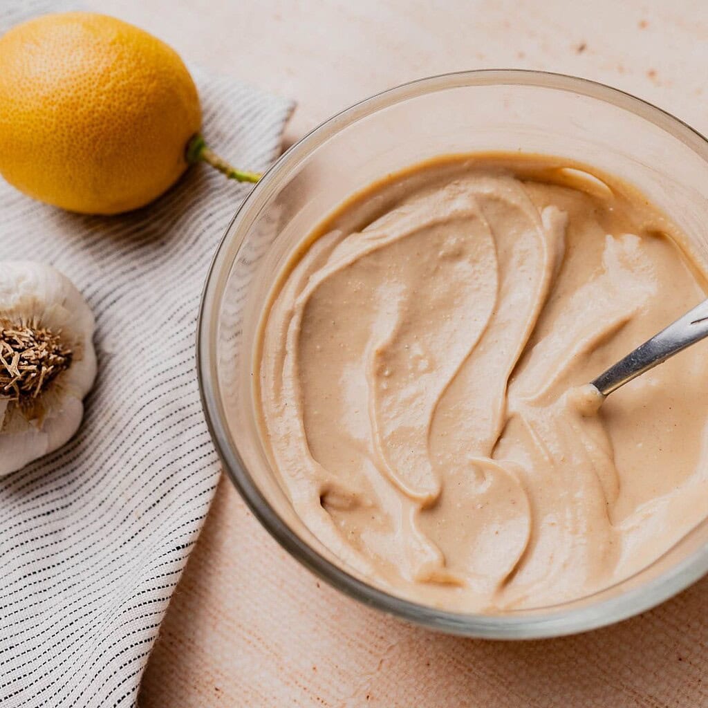 A glass bowl of creamy tahini sauce with a spoon, next to a lemon, a garlic bulb, and a striped cloth on a light surface—perfect for drizzling this savory tahini sauce over your favorite dishes.