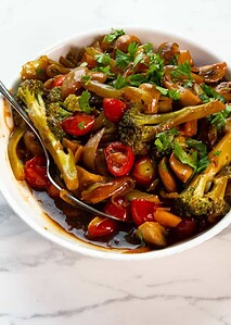 A white bowl filled with stir-fried vegetables including broccoli, mushrooms, onions, and cherry tomatoes, garnished with fresh herbs, on a marble surface.