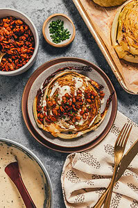 A roasted cabbage steak on a plate topped with creamy sauce, crispy lentils, and chopped chives, surrounded by bowls of sauce, lentils, and chives on a gray surface.