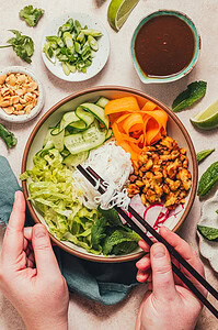 A person holds a bowl of rice noodles, lettuce, cucumber, carrots, radish, and cooked tofu with chopsticks, surrounded by herbs, peanuts, sliced green onions, lime, and a bowl of sauce.