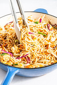 A blue skillet filled with stir-fried noodles, shredded vegetables, and scrambled eggs, with tongs lifting a portion.