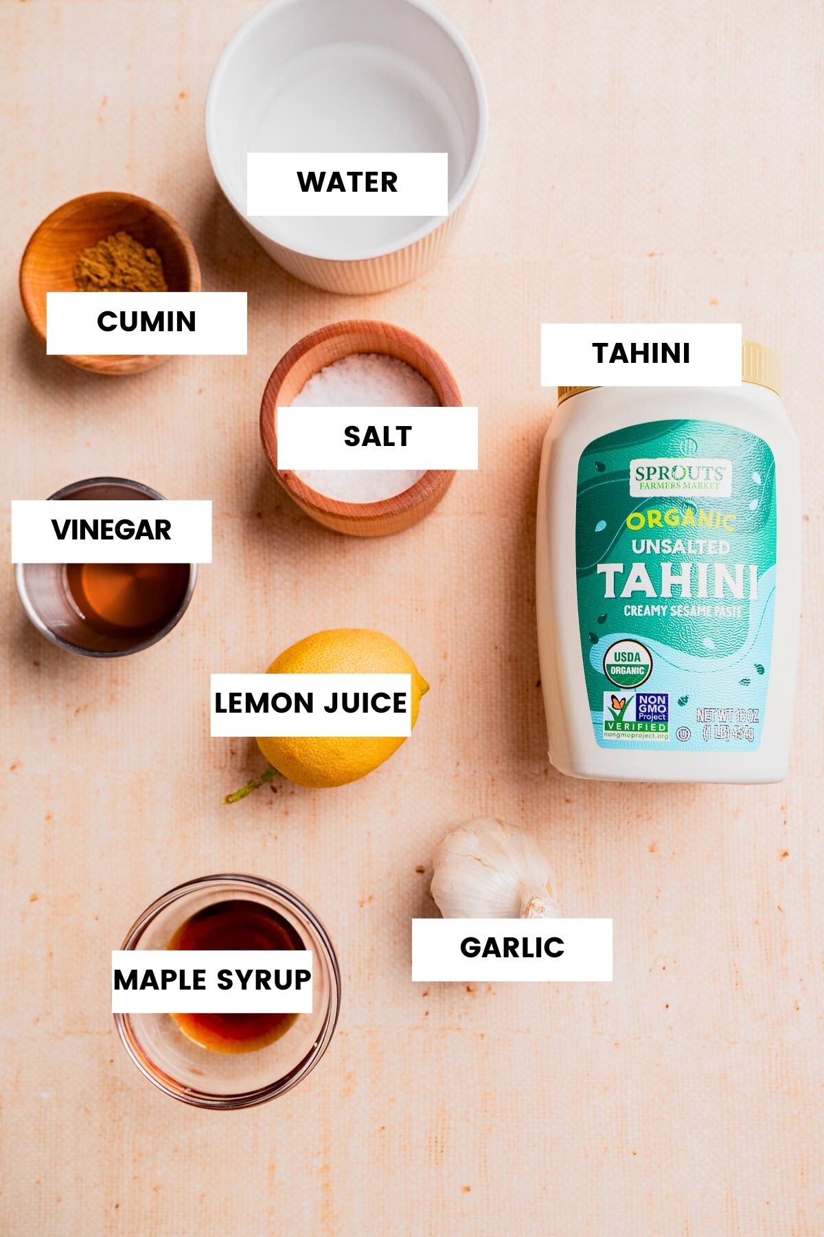 Labeled ingredients for Tahini sauce are arranged on a light surface, including cumin, water, salt, tahini, vinegar, lemon juice, maple syrup, and garlic.