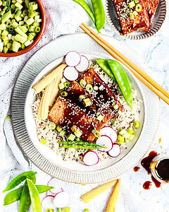 A bowl of rice topped with glazed tofu, sliced radishes, baby corn, and snow peas, garnished with sesame seeds and chopped green onions. Chopsticks and extra vegetables are nearby.