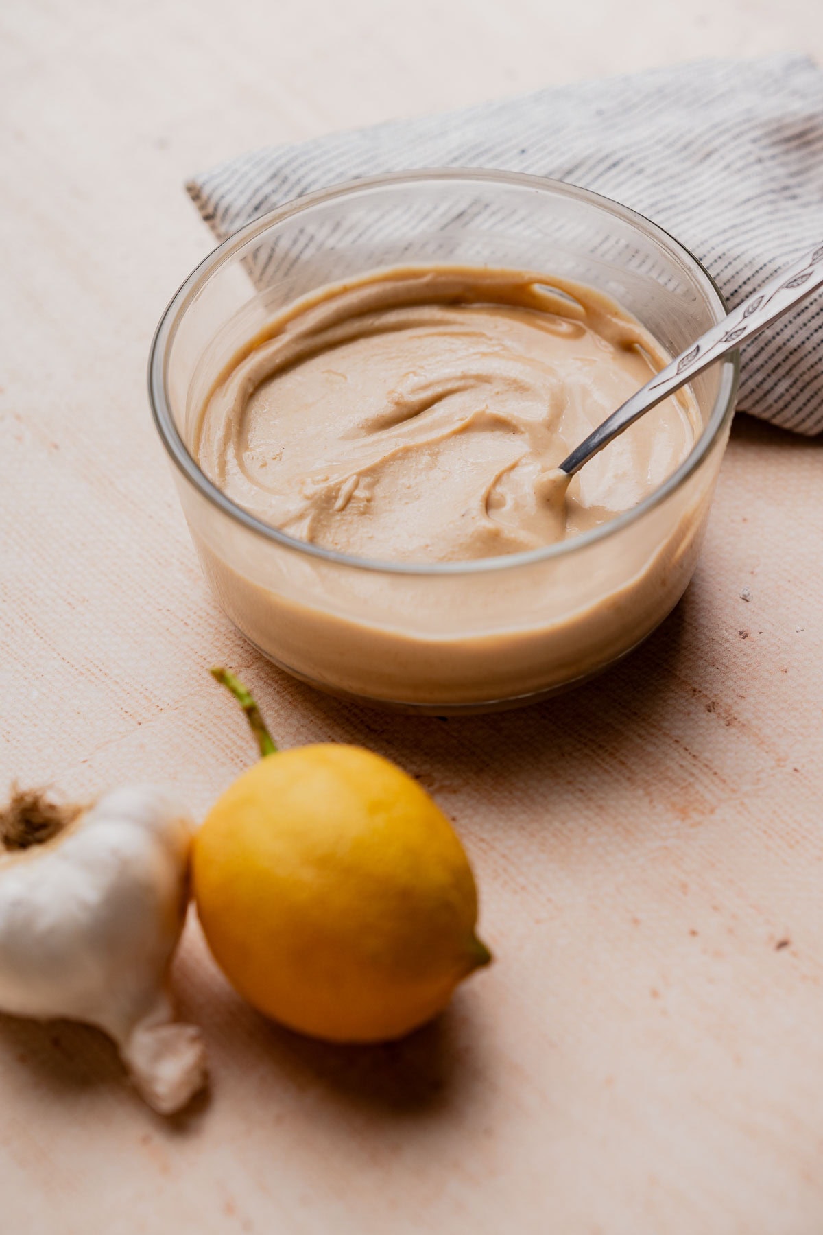 A glass bowl of creamy tahini sauce with a spoon, placed next to a lemon and a garlic bulb on a light surface with a striped cloth in the background.