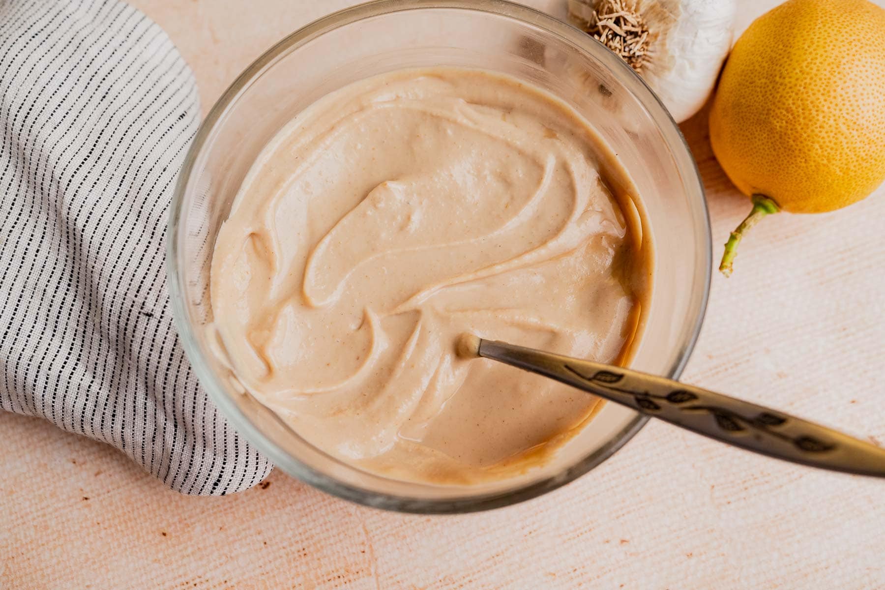 A glass bowl filled with creamy tahini sauce and a spoon, next to a lemon, a garlic bulb, and a striped cloth on a light surface.