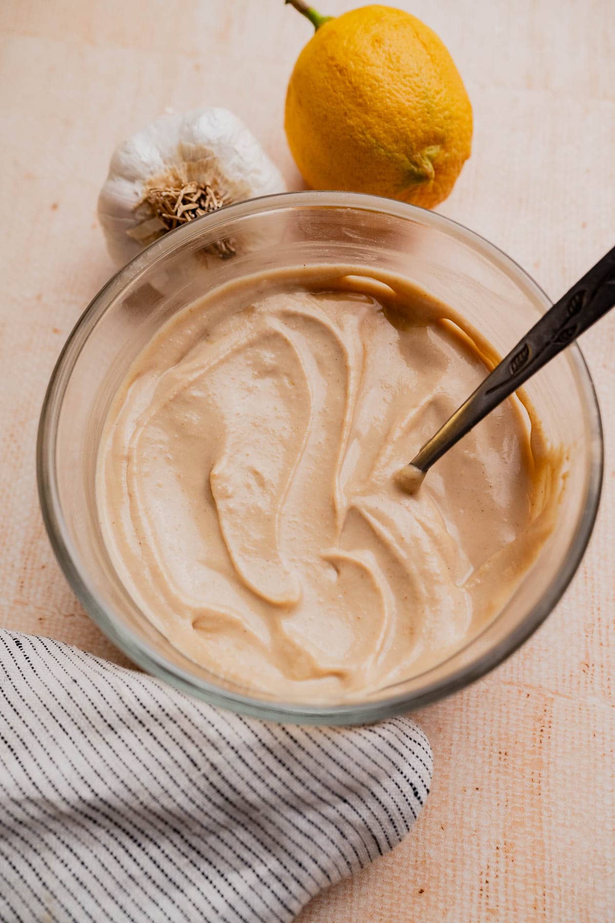 A glass bowl filled with creamy tahini sauce, with a metal spoon inside. Next to the bowl sit a whole lemon and garlic bulb, on a light surface topped with a striped cloth—perfect for drizzling this classic tahini sauce over your favorite dishes.