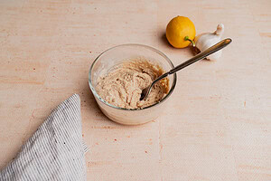 A glass bowl of creamy tahini sauce with a spoon, placed on a beige surface near a lemon, a garlic bulb, and a striped cloth.