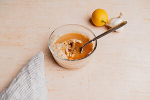 A glass bowl of creamy tahini sauce with a spoon, set beside a lemon and garlic on a light surface, with a striped cloth partially visible.