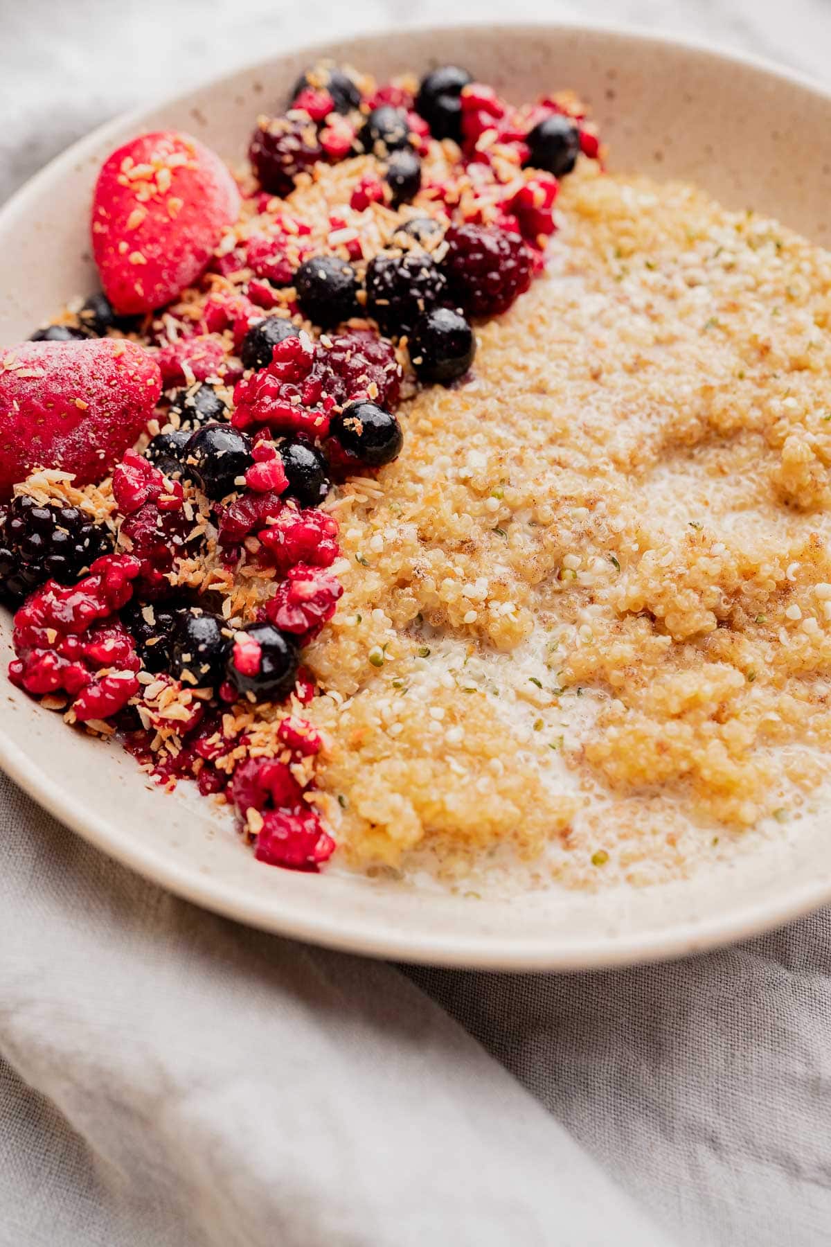 A bowl of quinoa breakfast porridge topped with mixed berries and shredded coconut, served on a beige plate.