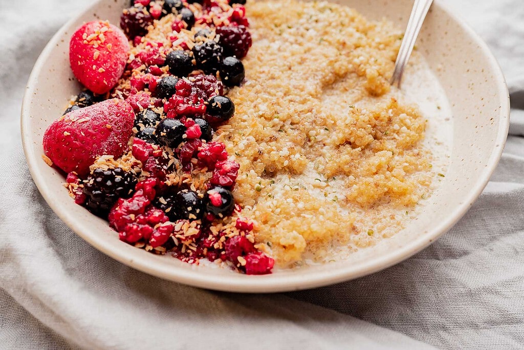 A bowl of quinoa breakfast porridge topped with strawberries, mixed berries, and shredded coconut, served with milk and a spoon.