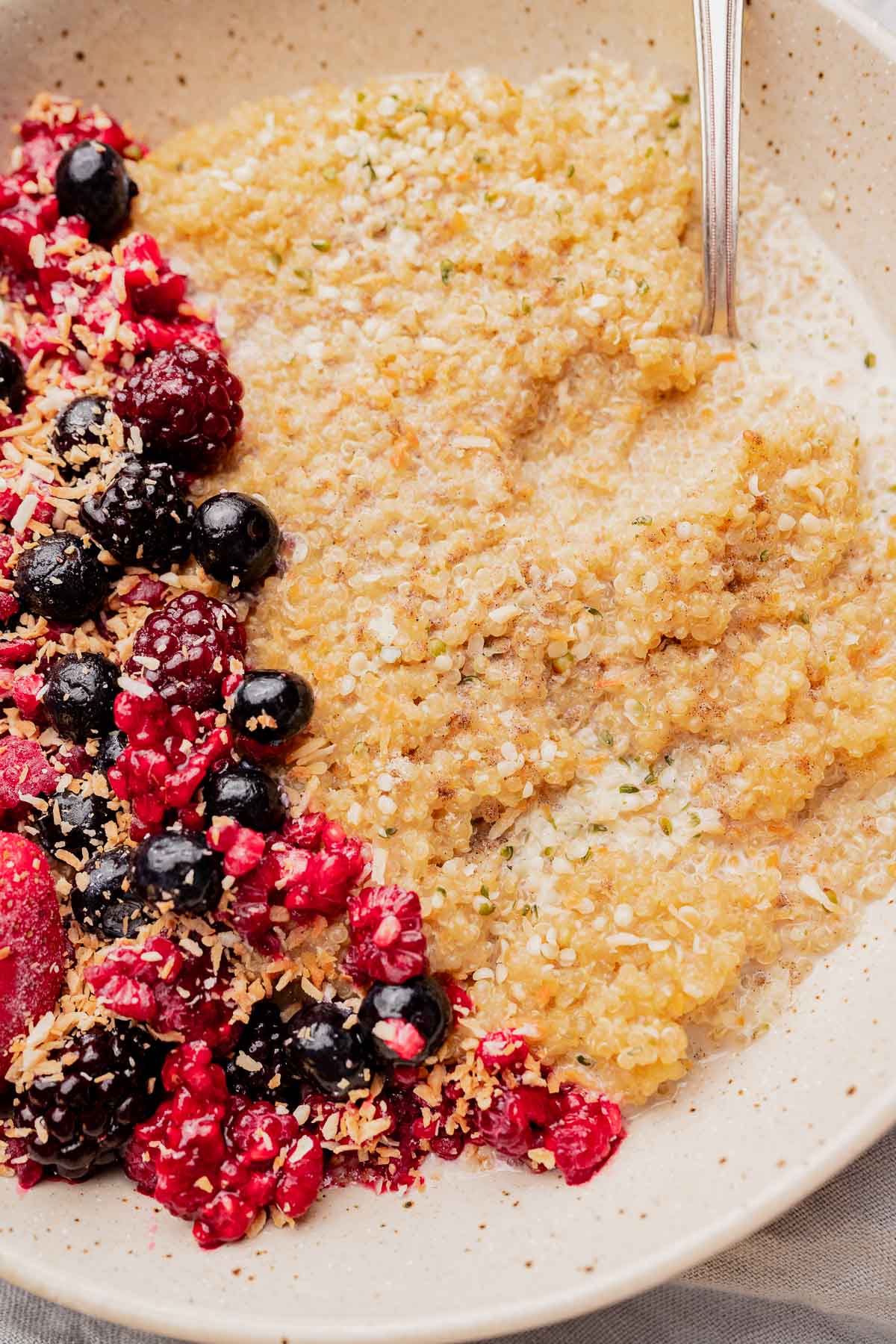 A bowl of quinoa breakfast porridge topped with mixed berries, shredded coconut, and hemp seeds, served with a spoon on the side.