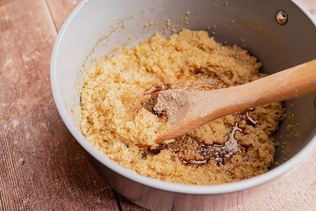 A saucepan with quinoa breakfast porridge, liquid, and seasoning being stirred with a wooden spoon on a wooden surface.