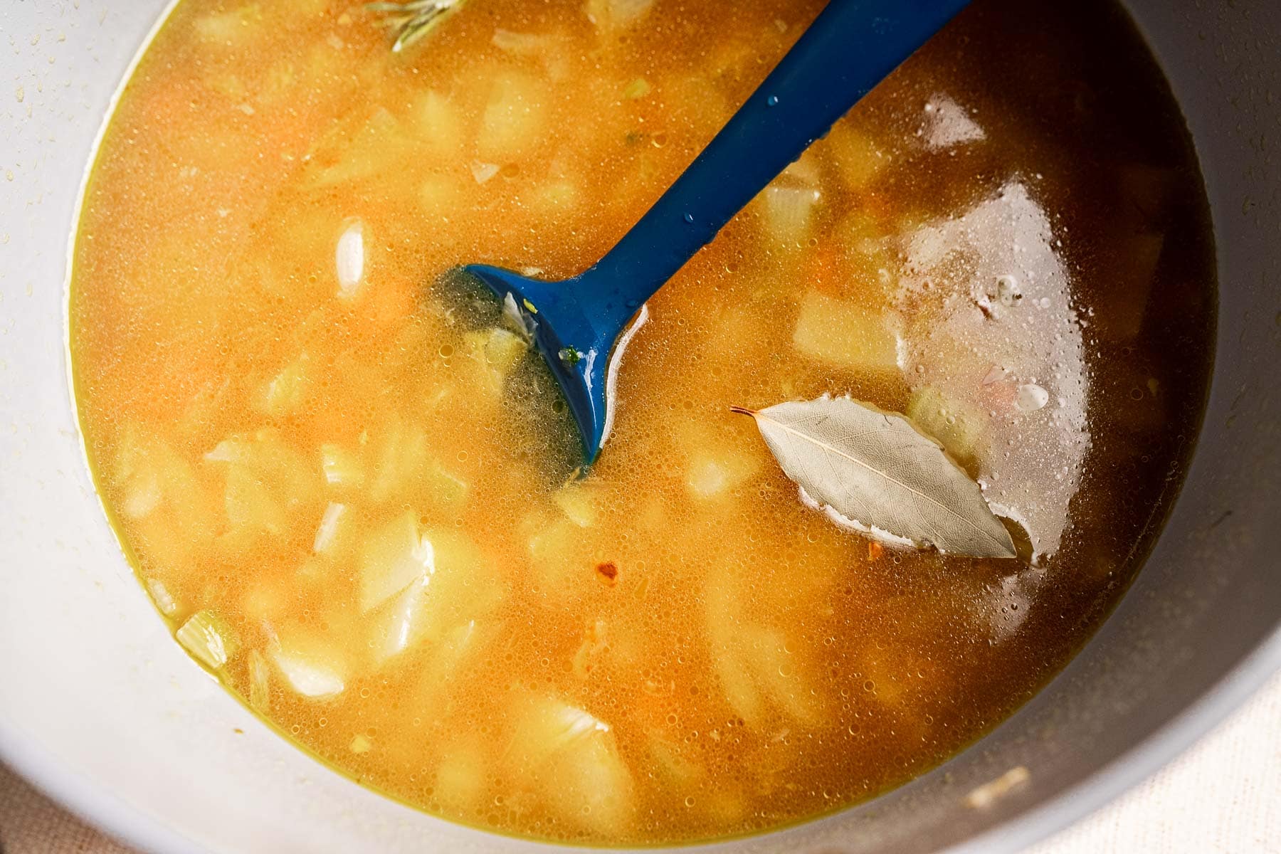 A bowl of Tuscan white bean soup with diced vegetables, a bay leaf, and a blue ladle partially submerged in the broth.