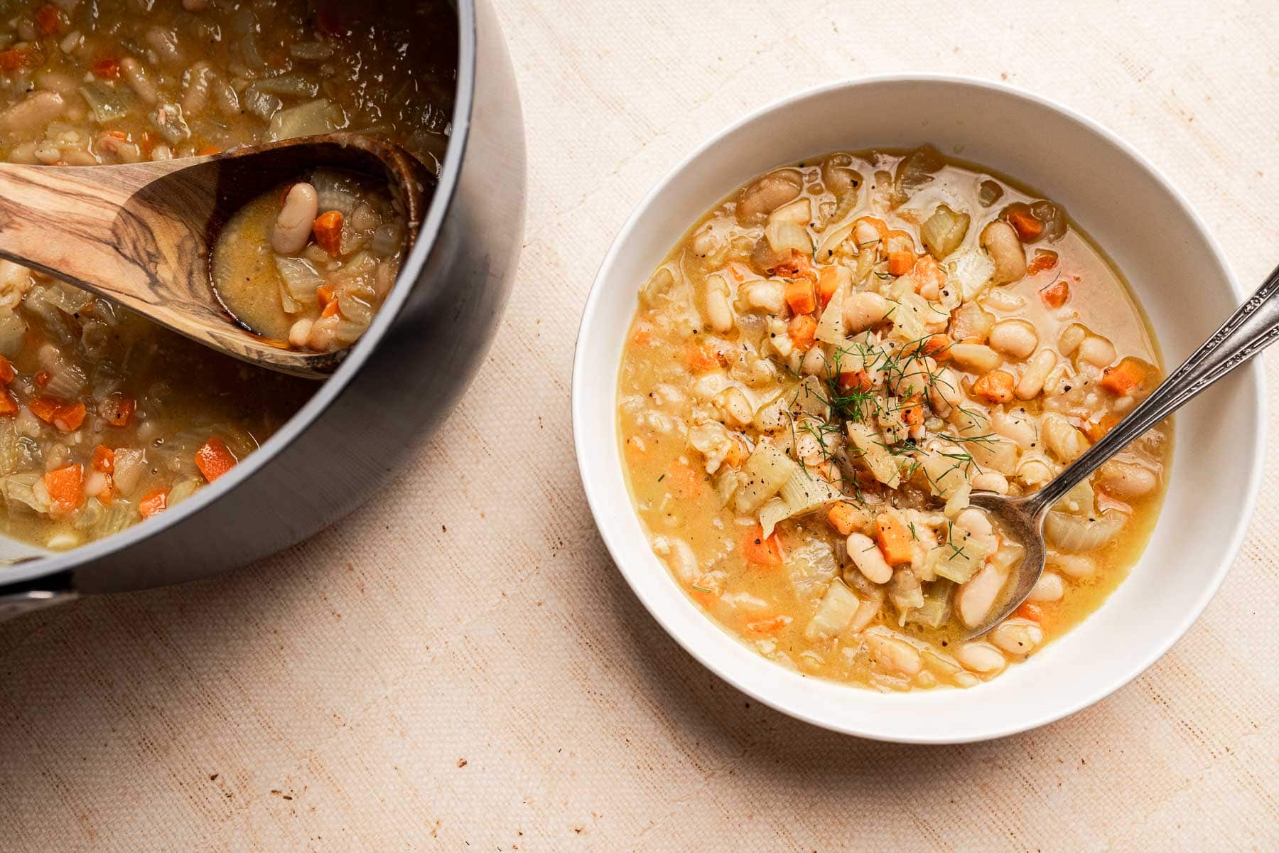 A bowl of Tuscan white bean soup with a spoon sits beside a pot of the same hearty vegetable soup and a wooden ladle, all arranged on a light-colored surface.