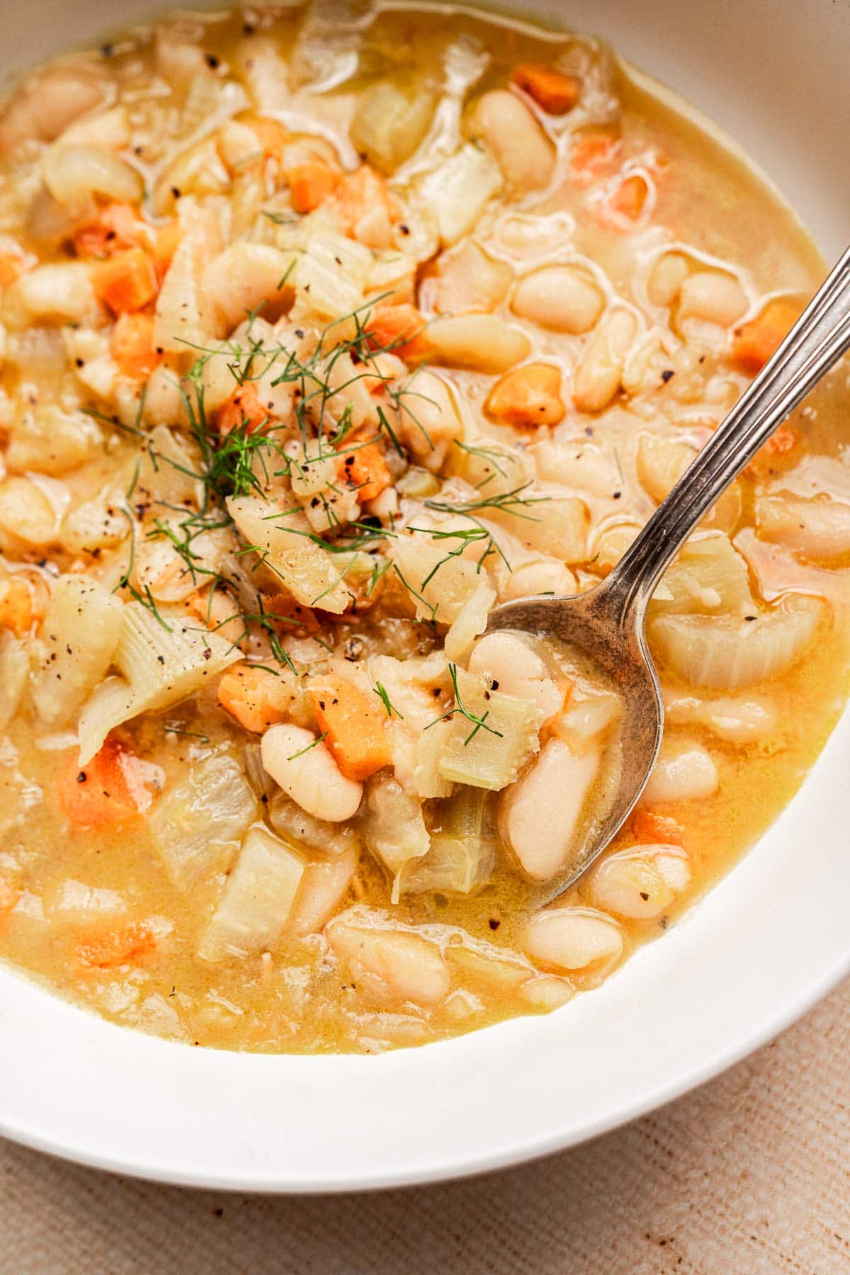 A bowl of Tuscan white bean soup with diced carrots, onions, and fresh herbs, shown with a spoon.