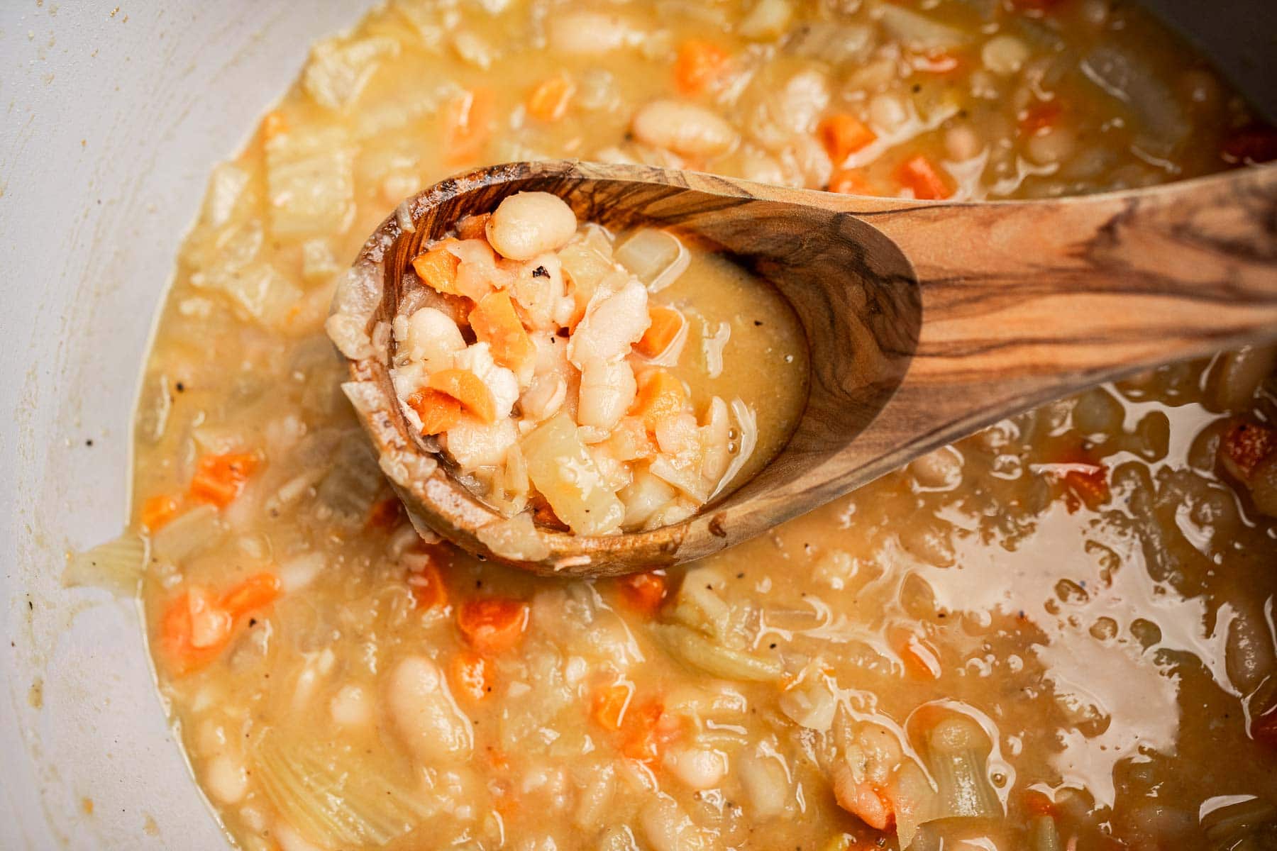 A wooden spoon holds a portion of Tuscan white bean soup, showcasing tender white beans, chopped carrots, and onions in a hearty, thick broth.