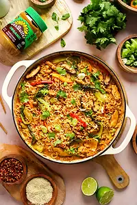 A pot of cooked ramen noodles with vegetables and herbs, surrounded by bowls of sliced green onions, cilantro, chili flakes, lime halves, sesame seeds, and a jar of peanut butter.