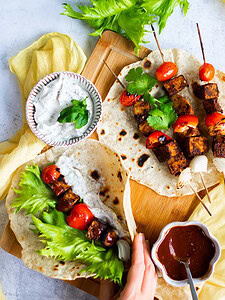 Two flatbreads topped with grilled vegetable and tofu skewers, lettuce, and sauces, served on a wooden board with a bowl of creamy dip and a bowl of reddish-brown sauce.