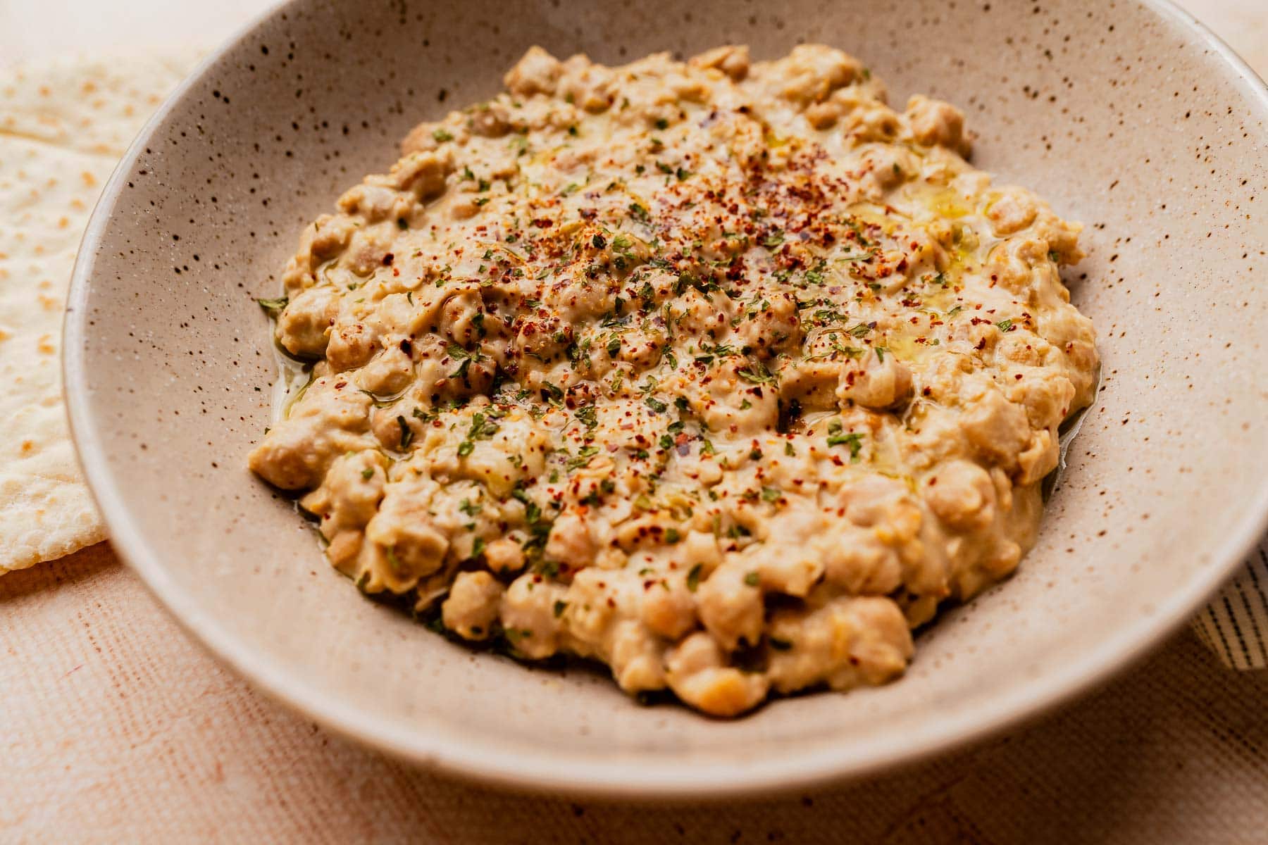 A close-up of a speckled bowl filled with creamy musabaha, a chickpea stew garnished with herbs and spices, on a light surface.