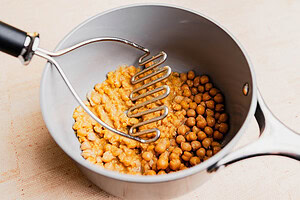 A potato masher mashing chickpeas in a silver saucepan for musabaha. Some chickpeas are whole while others are partially mashed.