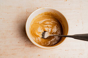 A metal fork rests in a white bowl containing a thick, light brown musabaha mixture on a textured beige surface.