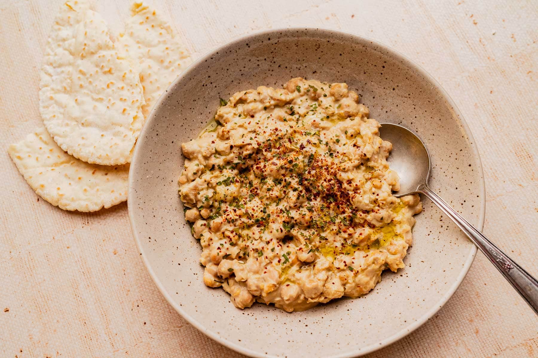 A bowl of chunky musabaha chickpea salad garnished with herbs and spices, served with a spoon and two pieces of flatbread on the side.