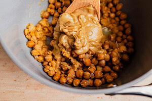 A wooden spoon stirs musabaha, blending chickpeas and tahini smoothly in a metal bowl.