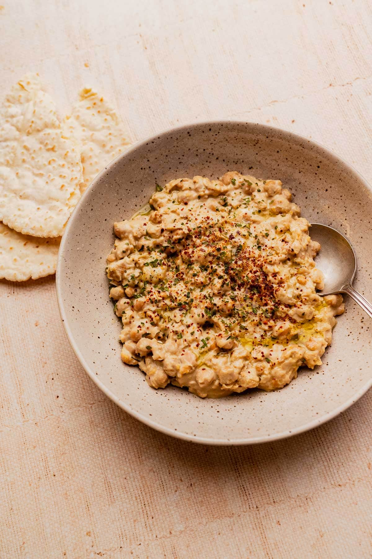 Bowl of creamy musabaha chickpea salad garnished with herbs and spices, served with flatbread on the side and a spoon in the bowl.