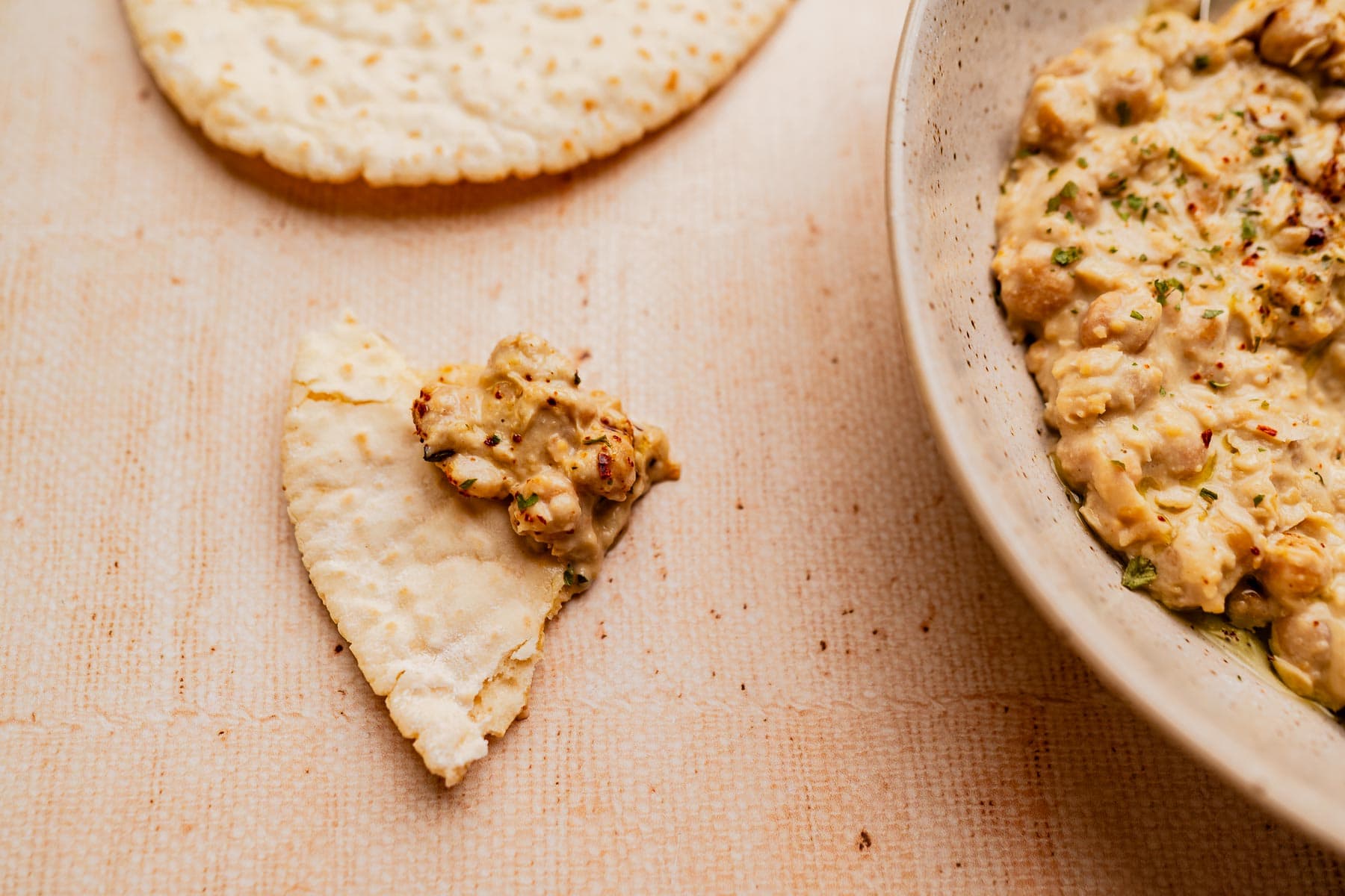 A piece of flatbread with a serving of creamy musabaha dip on it, next to a bowl containing more musabaha and another piece of flatbread on a beige surface.