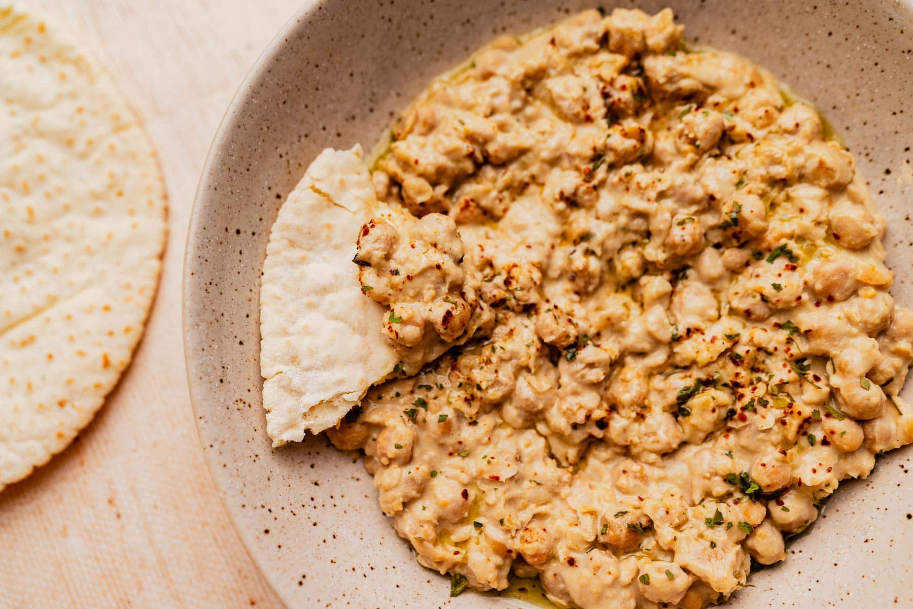 A bowl of musabaha chickpea salad garnished with herbs and spices, served with a piece of flatbread on the side.