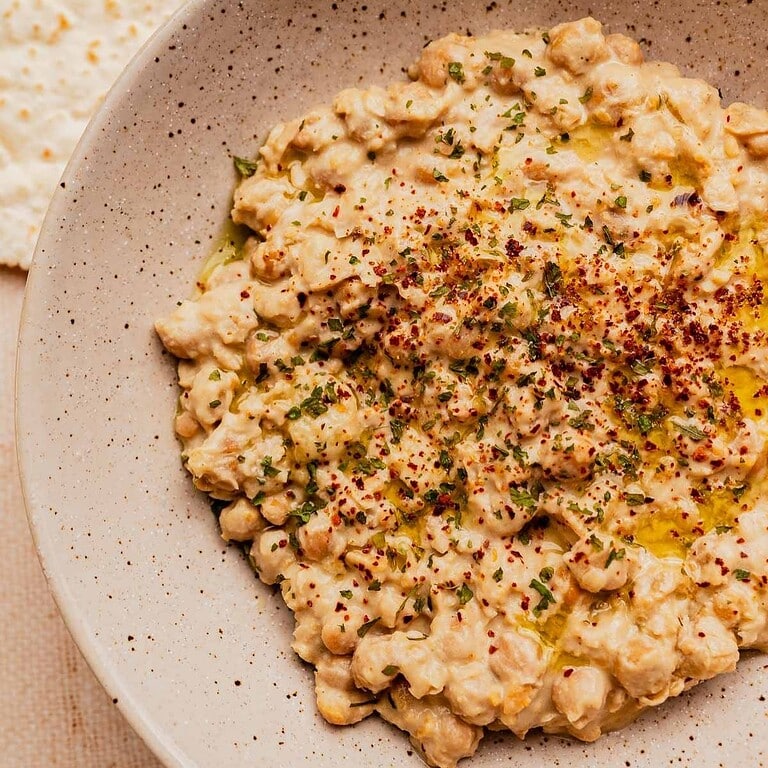 A bowl of musabaha-style chickpea salad with herbs, spices, and olive oil, served on a speckled plate.