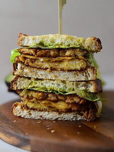 A stacked sandwich with tempeh, lettuce, and sauce on toasted bread, secured with a wooden pick, displayed on a wooden board.