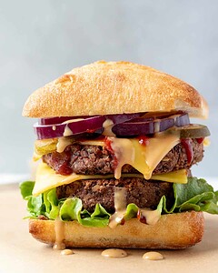 A double veggie burger with lettuce, cheese, pickles, red onion, and sauce on a ciabatta bun, with sauce dripping down.