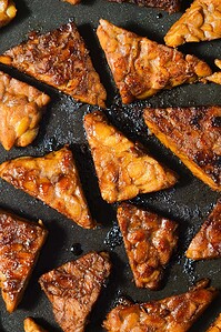 Triangular pieces of marinated tempeh are arranged on a dark pan, showing a glazed, browned surface after cooking.
