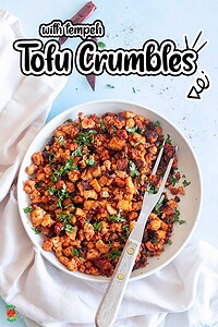 A white bowl filled with seasoned tofu crumbles, garnished with chopped herbs, sits on a white cloth; a fork rests inside the bowl.