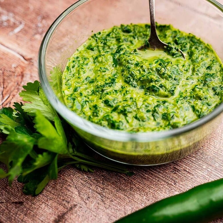 A glass bowl of green chimichurri sauce with a spoon, placed on a wooden surface next to fresh parsley and a green chili pepper, evokes the bold flavors of schug.