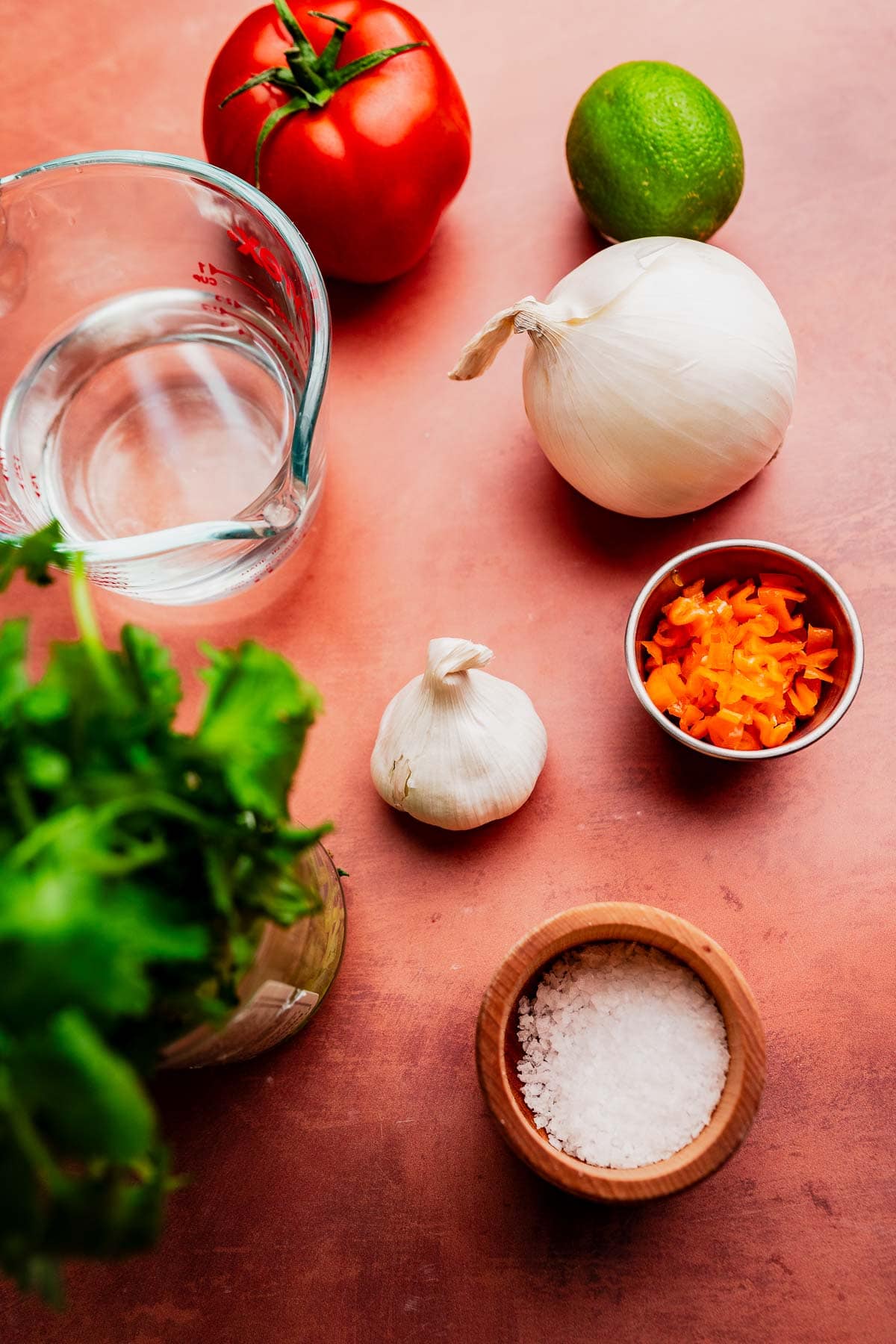 A tomato, white onion, lime, garlic bulb, chopped orange pepper, cilantro, a measuring cup of water, and a small bowl of salt sit on a brown surface—perfect ingredients for making fresh aji picante.