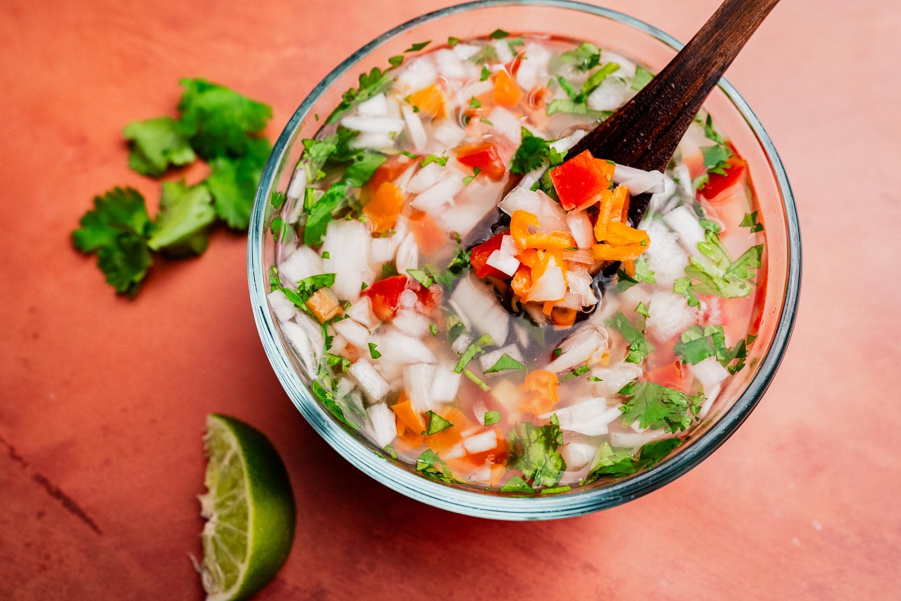 A glass bowl of chopped salsa with onions, tomatoes, cilantro, peppers, and a touch of aji picante, stirred with a wooden spoon; lime wedge and cilantro on the side.