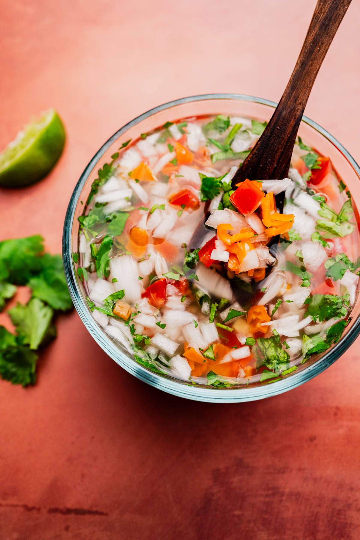 A glass bowl of chopped vegetables and herbs, including onions, tomatoes, and cilantro, perfect for aji picante, with a wooden spoon sits on a pink surface beside lime and cilantro leaves.