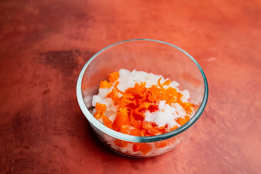 A glass bowl containing chopped white onions, chopped orange peppers, a heap of salt, and a hint of aji picante sits on a reddish-brown surface.