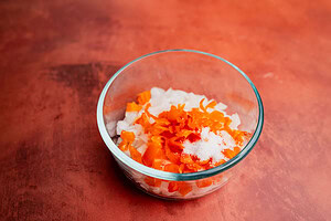 A glass bowl containing chopped white onions, chopped orange peppers, a heap of salt, and a hint of aji picante sits on a reddish-brown surface.
