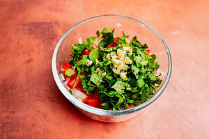 A glass bowl filled with chopped cilantro, diced tomatoes, onions, and minced garlic sits on a reddish surface—perfect ingredients for preparing a fresh aji picante.