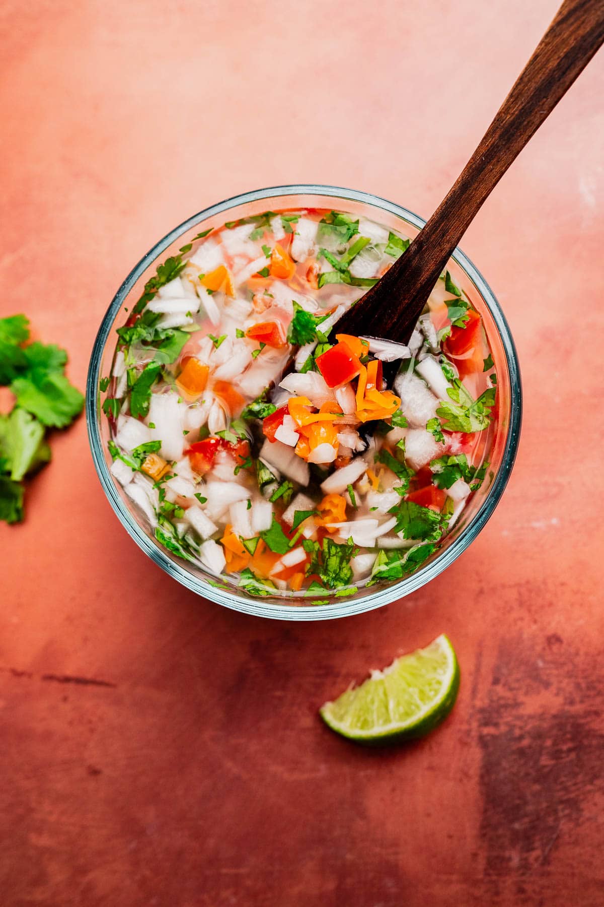 A bowl of freshly made salsa with chopped onions, tomatoes, cilantro, and peppers mixed with aji picante, stirred with a wooden spoon; a lime wedge and cilantro leaves are beside the bowl.