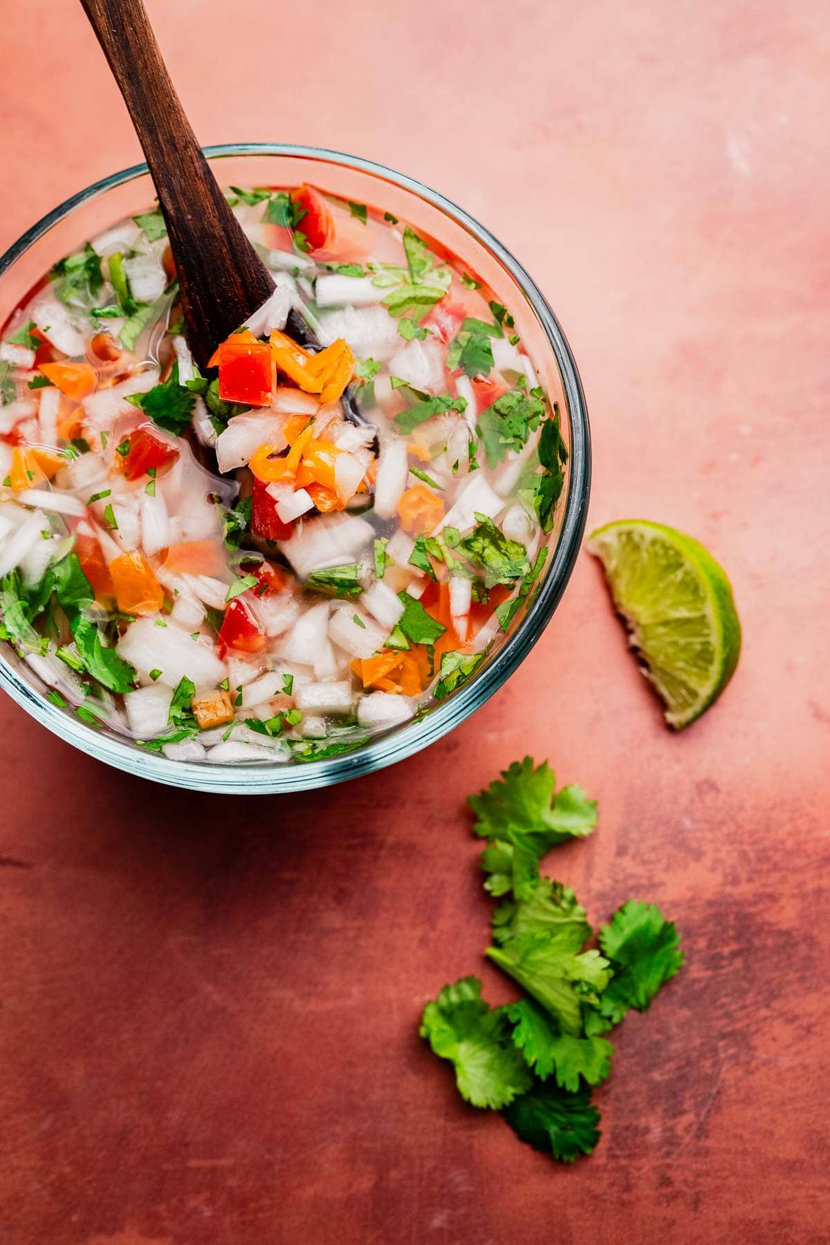 A glass bowl of chopped vegetables, including onions, tomatoes, cilantro, and carrots, with a wooden spoon. A lime wedge and cilantro leaves are on the table beside the bowl—perfect ingredients for preparing fresh aji picante.