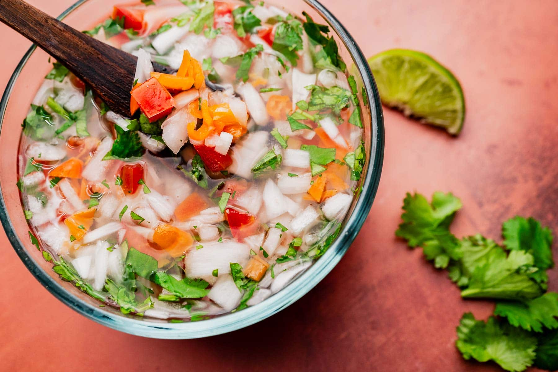 A glass bowl filled with chopped onions, tomatoes, peppers, and cilantro, mixed with aji picante. A wooden spoon sits inside, while a lime wedge and cilantro leaves are on the side.